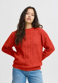 Pull en laine rouge avec un motif en torsades, à manches longues, col rond et ourlet côtelé. Porté avec un jean bleu clair sur un fond neutre.