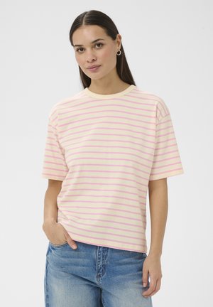 Woman with long dark hair wearing a cream and pink striped short-sleeve top and blue jeans, standing with one hand in pocket.