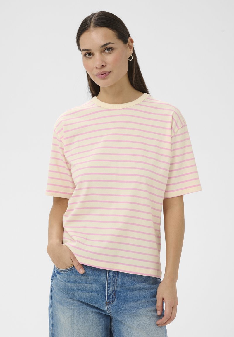 Žena s dlhými tmavými vlasmi, oblečená v krémovom a ružovom pruhovanom tričku s krátkym rukávom a modrých džínsoch, stojaca s jednou rukou v vrecku.