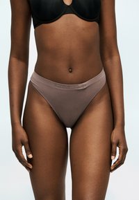 Γυναίκα που φοράει καφετί υψηλοκομμένα εσώρουχα Calvin Klein και μαύρο σουτιέν, στέκεται μπροστά σε απλό ανοιχτό φόντο.