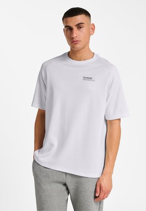 Junger Mann mit kurzen dunklen Haaren trägt ein lockeres weißes hummel-T-Shirt und graue Jogginghosen, steht mit einer Hand in der Tasche vor weißem Hintergrund.