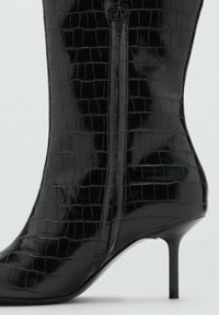 Bota negra hasta la rodilla en cuero sintético brillante con textura de cocodrilo, con cremallera lateral y punta afilada sobre un tacón de aguja.