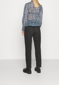 Donna con capelli biondi lisci, che indossa una blusa trasparente floreale blu navy, jeans dritti neri e scarpe con plateau nere, in piedi contro un muro bianco.