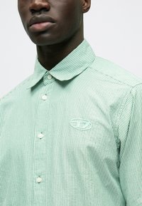 Cămașă cu nasturi cu dungi verzi și albe, guler ascuțit, cu un logo subtil pe pieptul stâng și material fin, texturat.
