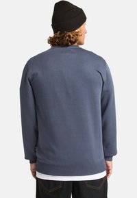Sudadera gris con puños y dobladillo acanalados. Presenta un cuello redondo y una textura suave. Usada sobre una camiseta blanca, exhibida desde la parte posterior.