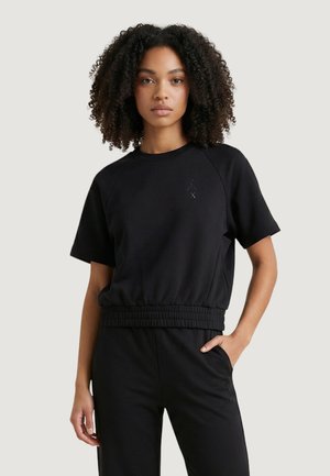 Mujer con cabello rizado, vestida con una camiseta negra de manga corta y pantalones negros a juego, de pie con una mano en el bolsillo.