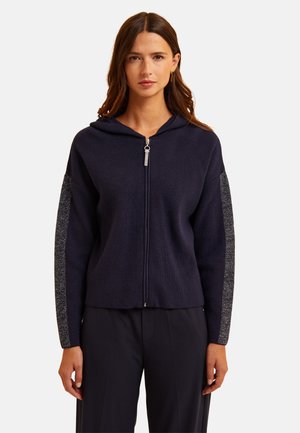 Frau in einer marineblauen Zip-Hoodie mit silbernen Streifendetails an den Ärmeln, kombiniert mit passenden marineblauen Hosen, vor einem einfarbigen Hintergrund stehend.