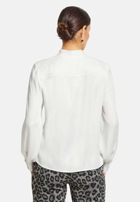 Weiße Bluse mit langen Ärmeln und einem leichten Glanz, mit einer Rückenpasse; kombiniert mit gemusterten grauen Leopardenhosen.