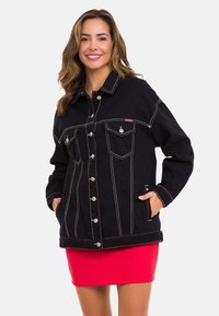 Schwarze Denimjacke mit kontrastierendem weißen Steppnähten, zwei Brusttaschen und silbernen Knöpfen. Über einem roten gerippten Minirock getragen.