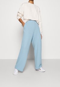 Helle blaue, weit geschnittene Hose aus strukturiertem Stoff, kombiniert mit einem lockeren cremefarbenen Sweatshirt. Das Model steht mit den Händen in den Taschen und trägt weiße Sneaker.