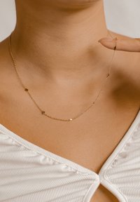 Collana delicata in oro con piccoli charm geometrici indossata al collo, tenuta da una mano con unghie curate in un top bianco testurizzato.