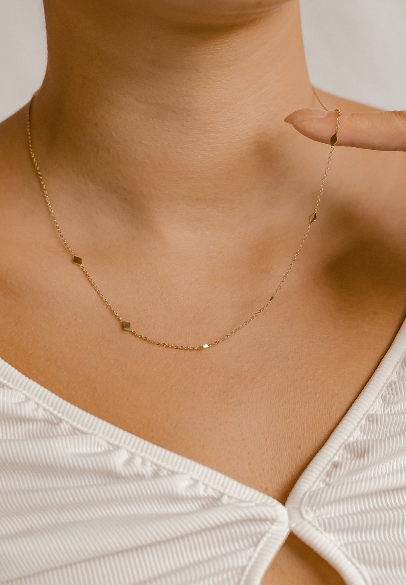 Collana delicata in oro con piccoli charm geometrici indossata al collo, tenuta da una mano con unghie curate in un top bianco testurizzato.