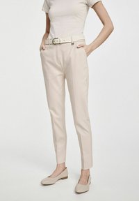 Beżowe spodnie o dopasowanym kroju z paskiem z okrągłą klamrą, prostym kroju, bocznymi kieszeniami i gładką fakturą; zestawione z beżowymi butami.