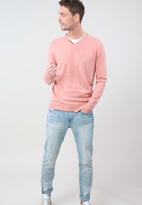 Sweater en tricot rose avec un décolleté en V et trois boutons, associé à un jean ajusté bleu clair et des baskets blanches. Texture de tissu lisse.