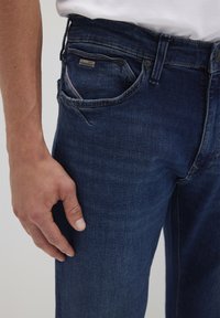 Jeans in denim blu scuro con taglio dritto, dotati di tasche anteriori, un bottone in metallo e una patch in pelle con logo sulla vita.