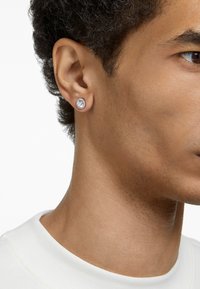 Brincos de pressão arredondados com uma pedra transparente embutida em um fecho prateado, usados na orelha esquerda de uma pessoa com cabelo escuro e cacheado.