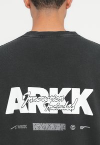 Čierna mikina s rebrovaným výstrihom, s veľkým bielym potlačou "ARKK" a nápisom "Inovácia zahrnutá" na chrbte, s ďalším textom nižšie.
