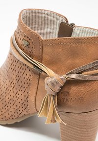 Gros plan sur une bottine en daim beige avec des côtés perforés, un talon empilé et un pompon en cuir décoratif noué à l'arrière du col.