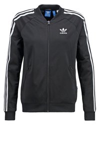 Zwarte polyester sportjas met ritssluiting aan de voorkant, ribgebreide manchetten en drie witte strepen langs elke mouw. Adidas-logo op de linkerborst.