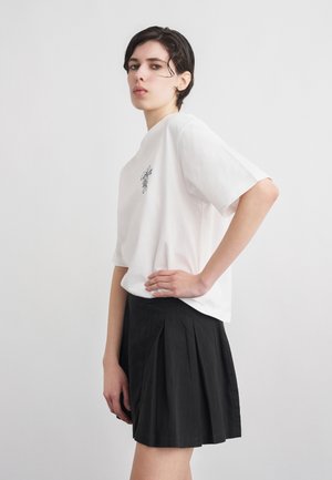 Νεαρός με κοντά σκούρα μαλλιά φοράει άσπρο φαρδύ μπλουζάκι με στάμπα και μαύρη πλισέ φούστα, ποζάροντας πλαγίως με το χέρι στη μέση.