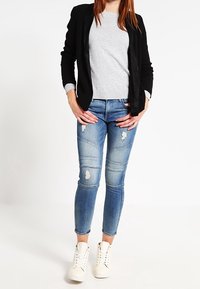 Jeans skinny de denim azul con detalles desgastados, combinados con un suéter gris y un blazer negro, terminando con zapatillas blancas de caña alta.