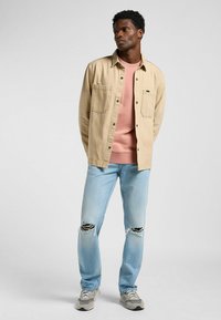Chaqueta beige de botones sobre un sudadera rosa, combinada con unos vaqueros claros desgastados y zapatillas grises. Fondo liso.
