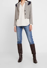 Blazer court à carreaux avec des accents bleu marine, associé à un chemisier léger, un jean bleu et des bottes texturées marron foncé jusqu'aux genoux.