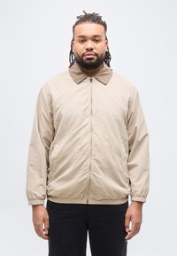 Άνδρας με πλεξούδες στα μαλλιά και γένια, φοράει μπεζ ζακέτα με φερμουάρ και μαύρο παντελόνι, στέκεται μπροστά σε απλό ανοιχτό γκρι φόντο.