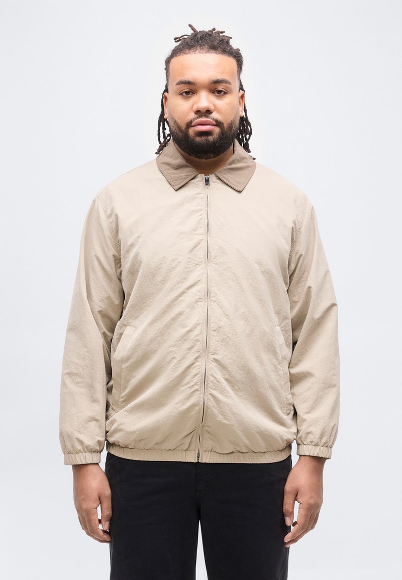 Άνδρας με πλεξούδες στα μαλλιά και γένια, φοράει μπεζ ζακέτα με φερμουάρ και μαύρο παντελόνι, στέκεται μπροστά σε απλό ανοιχτό γκρι φόντο.