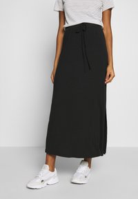 Jupe longue noire en tissu doux, dotée d'une taille à cordon, de fentes latérales, portée avec des baskets blanches et un t-shirt rayé.