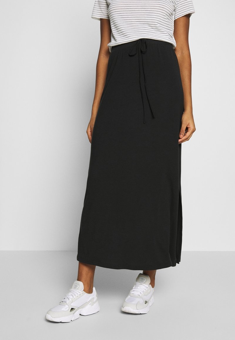 Jupe longue noire en tissu doux, dotée d'une taille à cordon, de fentes latérales, portée avec des baskets blanches et un t-shirt rayé.