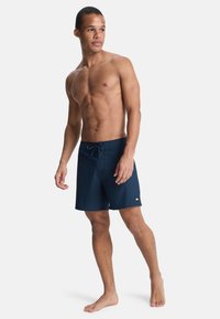 Σκούρο μπλε μαγιό με κλείσιμο με κορδόνι, κατασκευασμένο από ελαφρύ υλικό. Διαθέτει μια μικρή λεπτομέρεια λογότυπου στη κάτω αριστερή πλευρά.