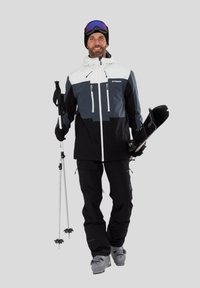 Skibekleidung mit einem farbigen Blockdesign in Schwarz, Grau und Weiß, ausgestattet mit Reißverschlüssen, Taschen und Skistöcken. Modell trägt Skischuhe.