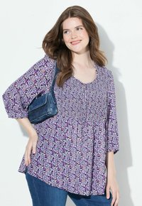 Bluse mit floralem Muster in Lila, mit einem gesmokten Oberteil und ¾-Ärmeln. Kombiniert mit einer denimbraunen Schultertasche und blauen Jeans.