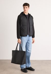 Schwarze gepolsterte Jacke mit Kapuze über einem schwarzen Shirt, kombiniert mit hellblauen Jeans und schwarzen flachen Sneakers, hält eine große schwarze Tragetasche.