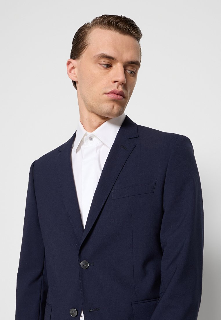 Mann mit nach hinten gegeltem Haar, der eine navyfarbene Anzugjacke und ein weißes Hemd trägt, schaut nach unten und zur Seite vor einem schlichten Hintergrund.