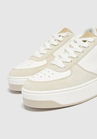 Beżowe i białe sneakersy z gładkiej skóry i zamszu, z perforowanym przodem, płaskimi sznurowadłami oraz grubą gumową podeszwą.