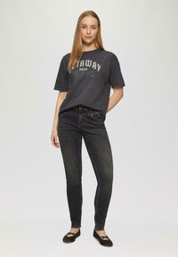 Czarnoszary t-shirt z grafiką, na którym widoczne są białe i turkusowe nadruki, zestawiony z czarnymi obcisłymi dżinsami i czarnymi płaskimi butami z krzyżującymi się paskami.