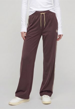 Personne portant un pantalon de survêtement marron à jambes larges avec des rayures crème et un cordon, un haut à manches longues blanc et des baskets blanches, debout.