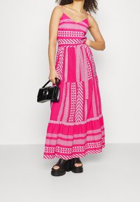 Robe maxi rose vif avec des motifs géométriques blancs, dotée de bretelles ajustables, d'un corsage ajusté et d'une jupe fluide. Sac à main noir et sandales épaisses.