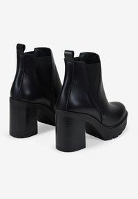 Un par de botas de cuero negro hasta el tobillo con tacones altos gruesos y paneles laterales elásticos sobre un fondo blanco.