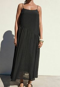 Robe longue noire avec fines bretelles, tissu texturé et silhouette fluide. Des bracelets décoratifs au poignet ajoutent une touche élégante.