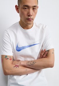 White cotton t-shirt featuring a blue Nike Swoosh logo with horizontal stripes. Model with visible tattoos crossing arms.