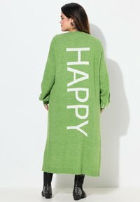 Cárdigan largo de punto verde con grandes letras blancas que dicen "HAPPY" en la parte posterior, con mangas sueltas y una textura suave. Botines negros con tachuelas exhibidos.