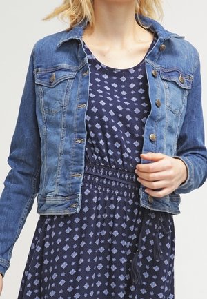 Woman wearing a blue patterned dress with a smocked waist and a buttoned blue denim jacket, with one hand adjusting the jacket.