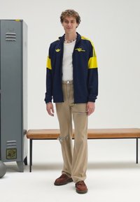 Laivastonsininen takki keltaisin yksityiskohdin ja logolla, valkoinen paita, beigenväriset revityt housut, ruskeat kengät, seisoo harmaan kaapin ja penkin vieressä.