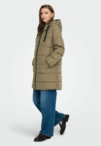 Maslinasto zeleni kaput s kapuljačom, s prednjim zatvaračem, crnim vezicama i bočnim džepovima. Uparen s širokim plavim trapericama i crnim cipelama.