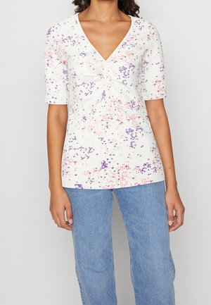 Frau trägt ein weißes Kurzarmoberteil mit kleinen rosa und violetten Blumenmustern und blaue Jeans, steht vor einem einfarbigen Hintergrund.