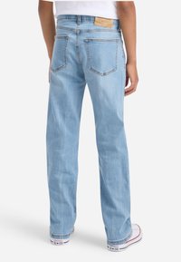 Cilvēks, kas ģērbjas gaiši zilos taisnās kājas džinsos un baltos Converse apavos, redzams no aizmugures pret vienkāršu fona.