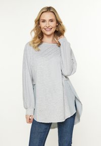 Lys grå langærmet top med en afslappet pasform, der har en blød tekstur, sideslids og lagdelt kant for ekstra detaljer. Sammen med blå jeans.